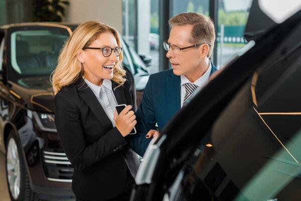 businessman and smiling businesswoman with smartphone choosing new car in dealership salon
