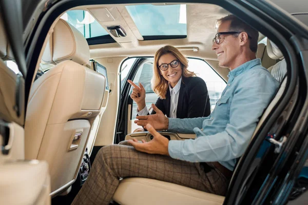 快乐的成年男子和女汽车经销商坐在豪华轿车后座的侧面视图和交谈 — 图库照片