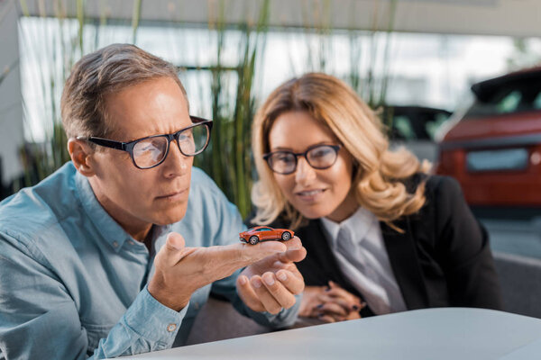 thoughtful adult customer and smiling female car dealer looking at toy car in showroom