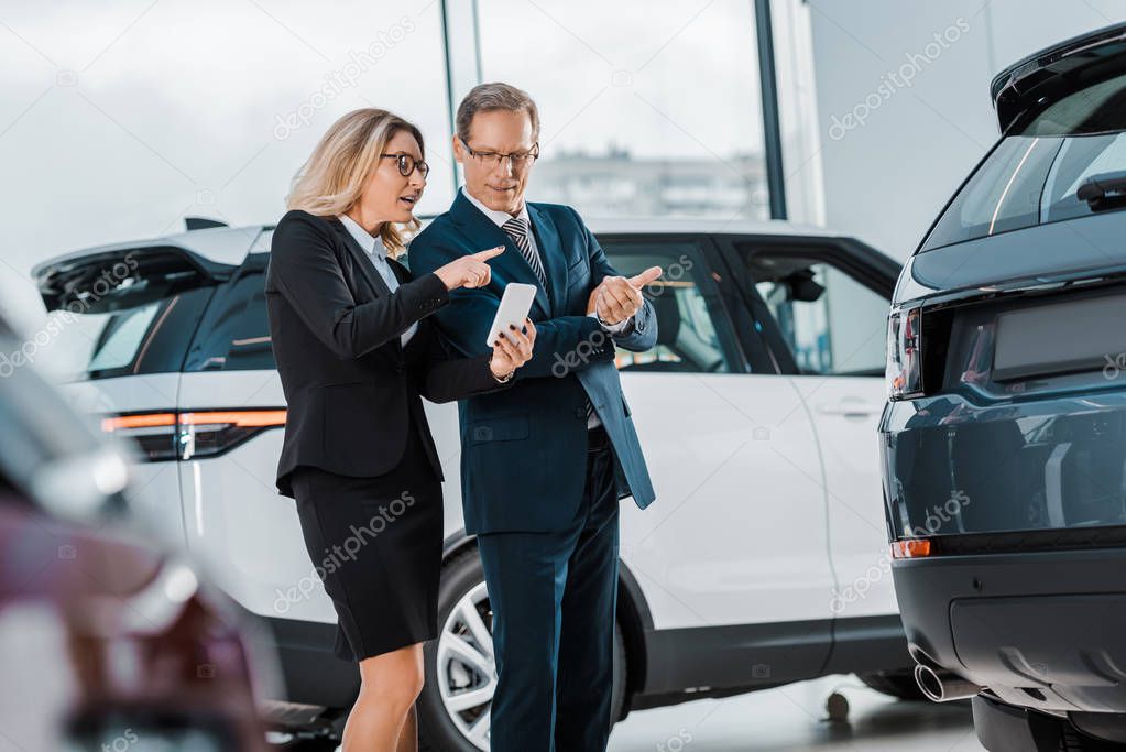 businessman and businesswoman with smartphone choosing new automobile in showroom