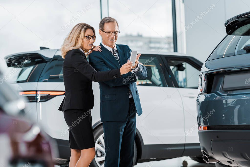 businessman and businesswoman with smartphone choosing new automobile in showroom
