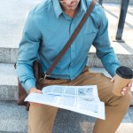 Cropped shot of businessman with coffee to go sitting on stairs and reading newspaper