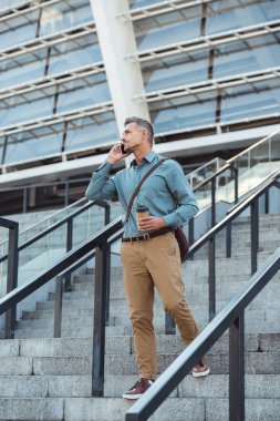 handsome man talking by smartphone and looking away while standing with coffee to go on stairs clipart