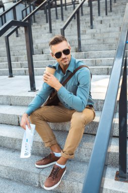 smiling man in sunglasses holding newspaper and drinking coffee from paper cup on stairs clipart