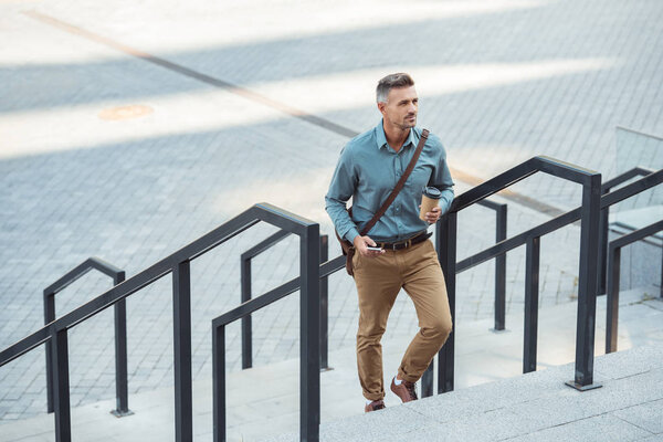 high angle view of man holding paper cup and using smartphone on stairs