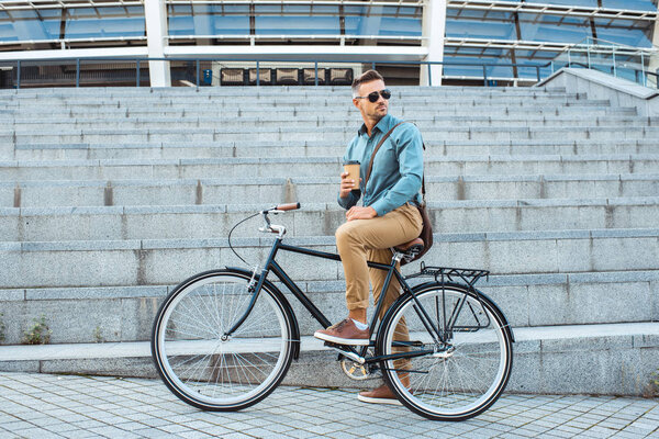 Красивый мужчина в солнечных очках сидит на велосипеде с кофе, чтобы пойти и оглянуться через плечо на улице
