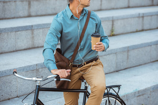 cropped shot of man in earphones holding paper cup and sitting on bicycle