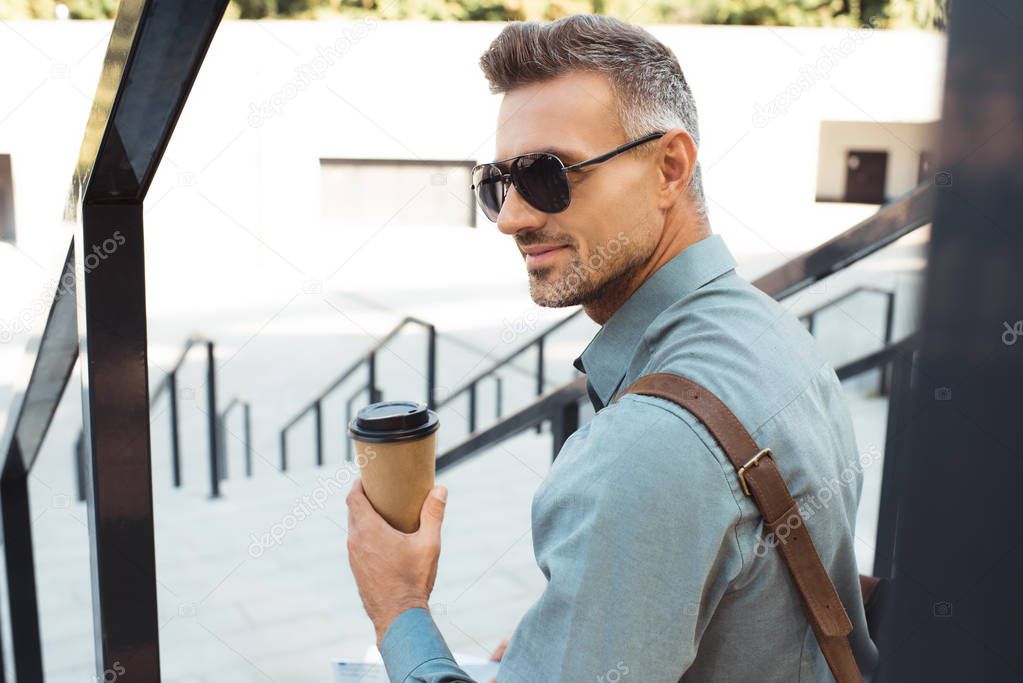 handsome smiling man in sunglasses holding coffee to go and sitting on stairs