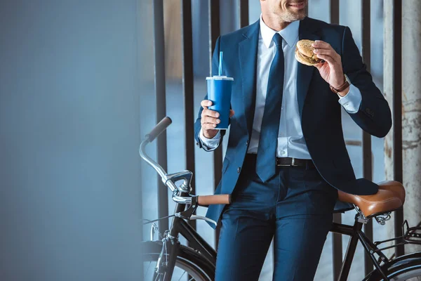 使い捨てカップとハンバーガーを押しながら自転車に座って笑顔の実業家のショットをトリミング — ストック写真