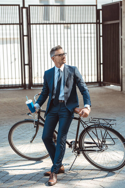 handsome smiling businessman holding paper cup and sitting on bicycle on street