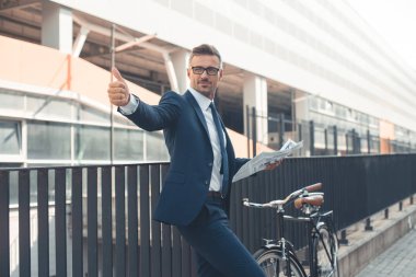 gazete tutan ve bisiklet için sokakta giderken başparmak gösterilmesini işadamı gülümseyen