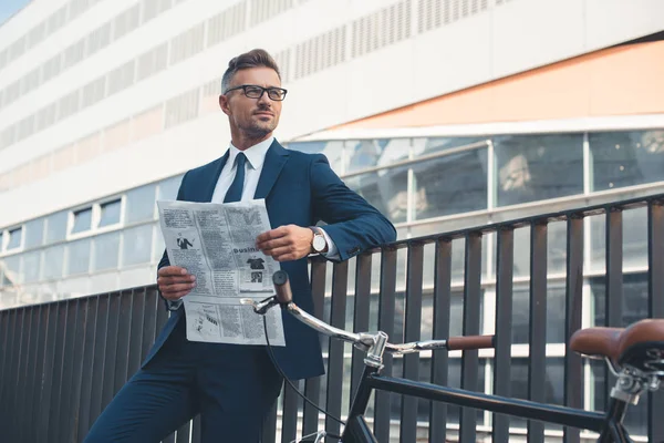 Businessman in suit and eyeglasses holding newspaper and looking away while standing with bike on street — Stock Photo, Image