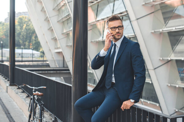 handsome businessman in suit and eyeglasses talking by smartphone and looking away while sitting on street