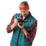 Fotografo maschio hipster sorridente in gilet di denim e camicia a scacchi guardando lo schermo della fotocamera isolato su bianco