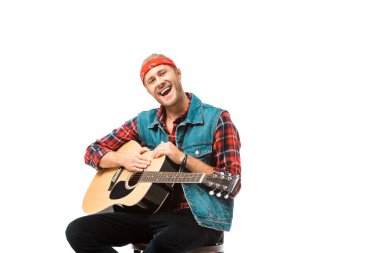 Akustik gitar üzerinde beyaz izole oturan denim yelek gülümseyen hipster erkekte