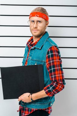 arrested hipster man in eyeglasses and headband holding empty prison board in front of police line up
