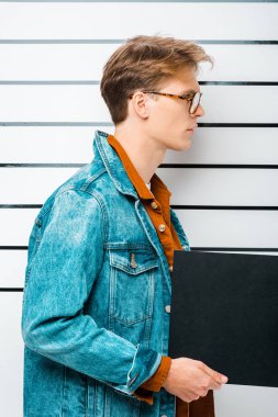 side view of arrested hipster man in eyeglasses holding empty prison board in front of police line up clipart