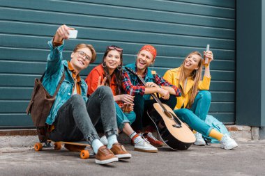 young friends with beer, skateboard and guitar taking selfie on smartphone clipart