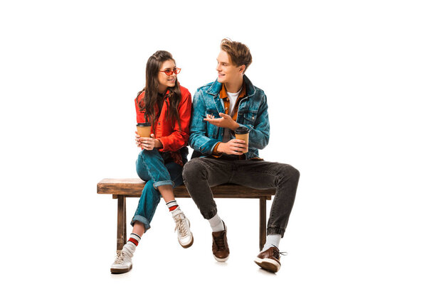 male hipster with paper cup of coffee pointing to girlfriend sitting on bench isolated on white