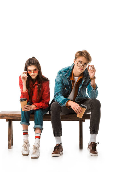fashionable hipster couple with coffee cups adjusting eyeglasses and sitting on bench isolated on white