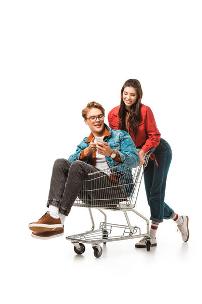young male hipster in shopping cart using smartphone while his girlfriend standing behind isolated on white