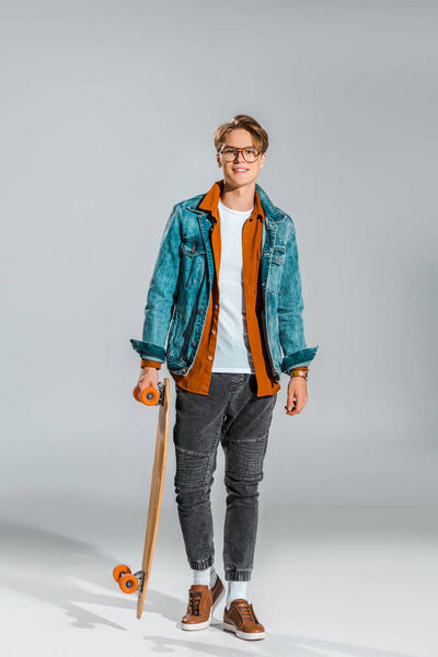 young hipster in denim jacket posing with skateboard on grey