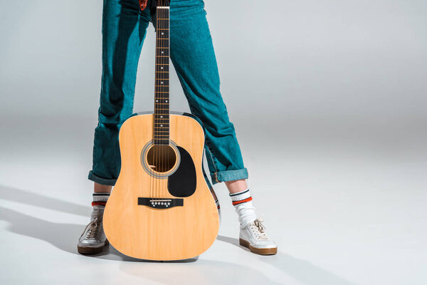 обрезанный вид девушки в джинсах с акустической гитарой на сером
