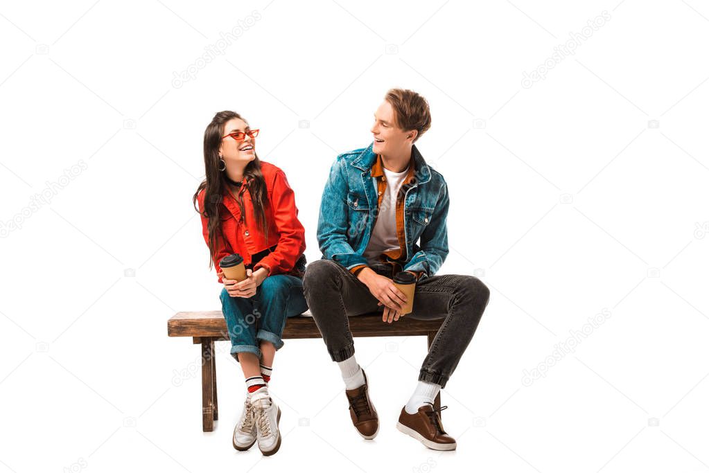 laughing hipster couple with disposable cups of coffee sitting on bench isolated on white