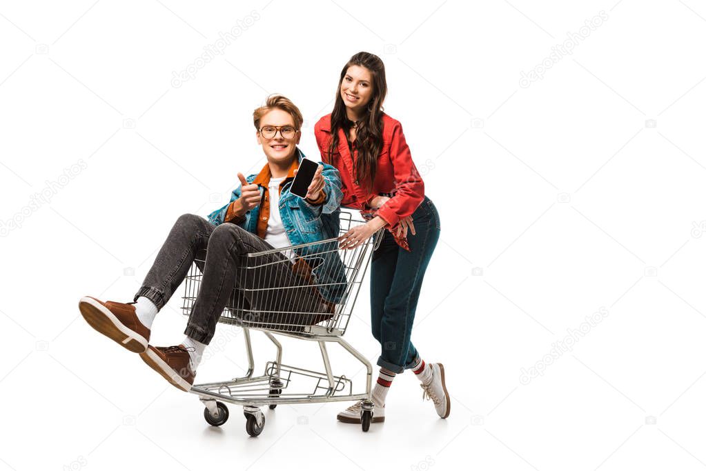 stylish male hipster in shopping cart showing smartphone with blank screen and doing thumb up while his girlfriend standing behind isolated on white