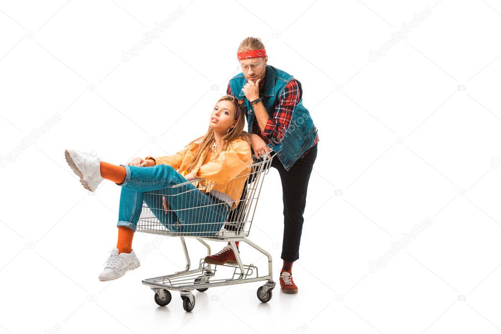 attractive hipster girl sitting in shopping cart while her boyfriend standing behind isolated on white
