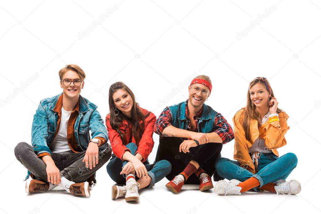 smiling hipster friends sitting on floor and looking at camera isolated on white