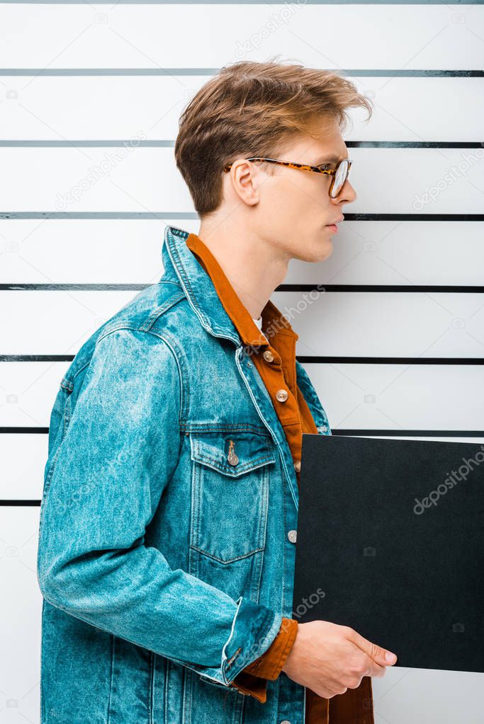 side view of arrested hipster man in eyeglasses holding empty prison board in front of police line up