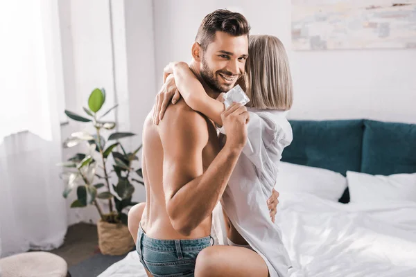 Smiling Young Man Holding Condom While Embracing Girlfriend Bedroom Looking — Stock Photo, Image