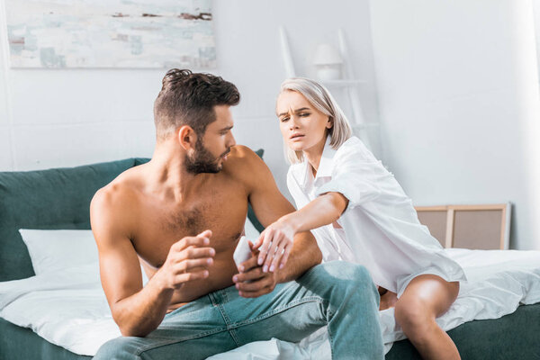 jealousy young man using smartphone of his angry girlfriend while she sitting on background in bedroom