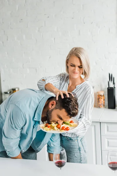 愤怒的女朋友粉碎男友脸到沙拉在盘子在厨房 — 图库照片