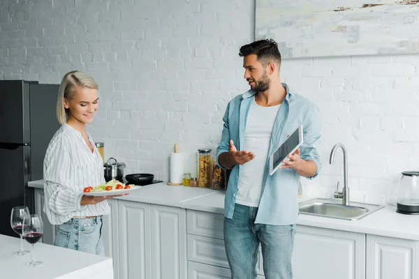 男友指着平板电脑微笑的女朋友与盘子在厨房 — 免费的图库照片