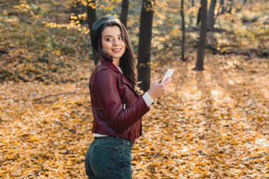 cheerful fashionable girl in beret and leather jacket holding smartphone and looking away outdoors 