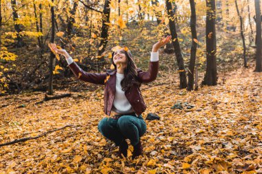 smiling attractive woman in stylish leather jacket having fun with yellow leaves in autumnal forest  clipart