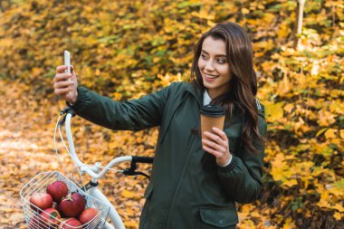 Smartphone cep telefonu ile sonbahar ormandaki selfie alarak tek kullanımlık kahve fincanı ile neşeli güzel kadın 