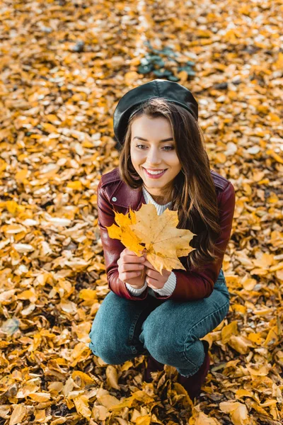 美丽的女模特在贝雷帽和皮革夹克坐在秋天公园黄色 — 图库照片