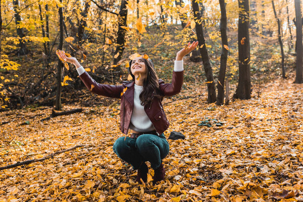 smiling attractive woman in stylish leather jacket having fun with yellow leaves in autumnal forest 