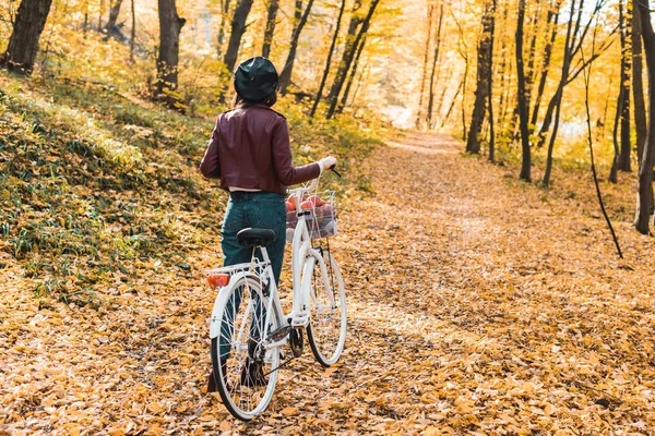 スタイリッシュな革のジャケット ベレー帽紅葉の森で自転車を運ぶ女性の背面図  — 無料ストックフォト