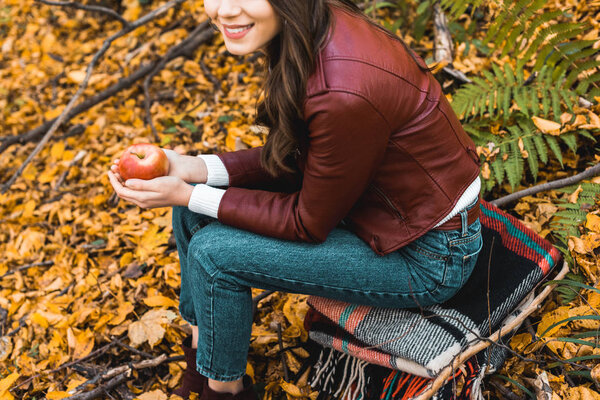 partial view of stylish girl in leather jacket sitting on blanket and holding red apple in autumnal forest