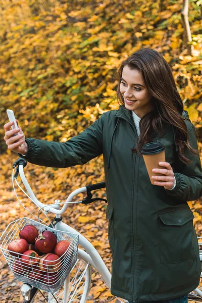 有吸引力的微笑的妇女与一次性咖啡杯拍摄自拍在秋季森林智能手机 — 免费的图库照片