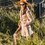 Modelo feminino elegante em óculos de sol, casaco de trincheira e chapéu posando perto da grama