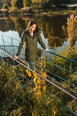 high angle view of young woman in jacket and yellow rubber boots posing near pond in park clipart