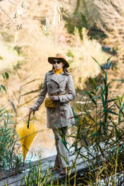young woman in sunglasses, trench coat and hat posing with yellow umbrella near pond in park clipart