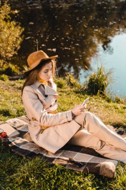 battaniye yakınındaki park su birikintisi üzerinde otururken smartphone kullanarak şapkalı şık genç kadın