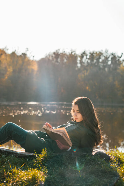 attractive girl laying on blanket and reading book near pond in park 