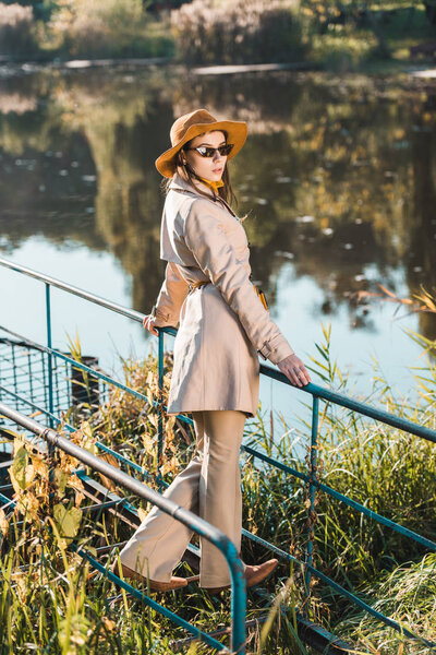 селективный фокус стильной женской модели в солнцезащитных очках, плаще и шляпе, позирующих возле пруда в парке

